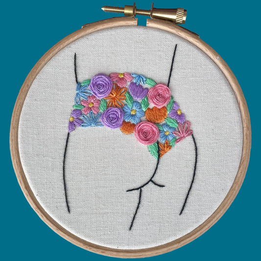 Embroidery hoop with a colourful floral knicker design on natural calico fabric against a teal background.