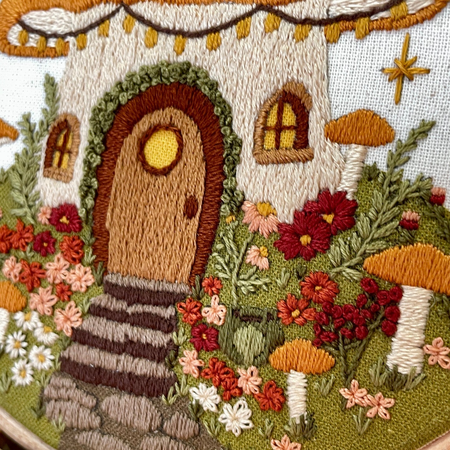 Embroidered design of a fairy house with a mushroom roof, surrounded by flowers and plants.