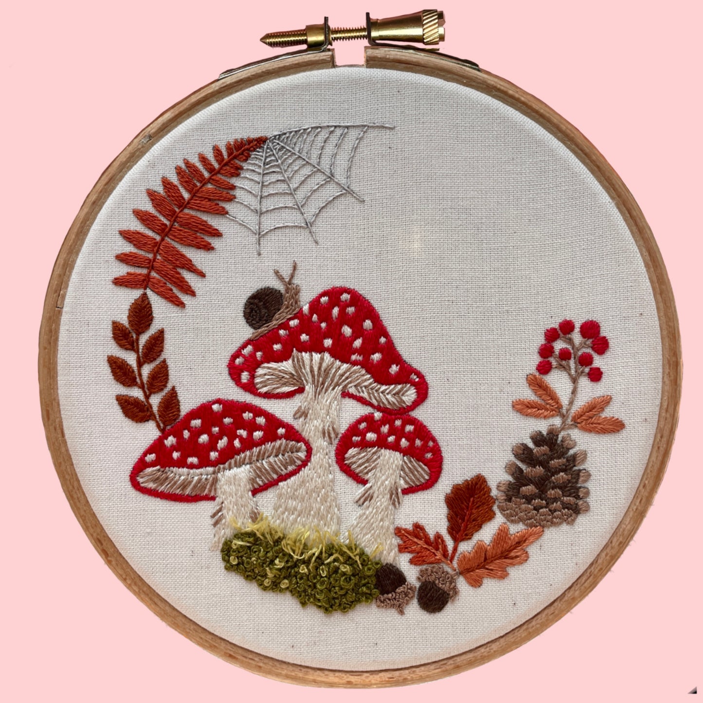 Embroidery kit depicting red mushrooms with white spots, accompanied by leaves and a spider web, all embroidered on a beige fabric hoop.