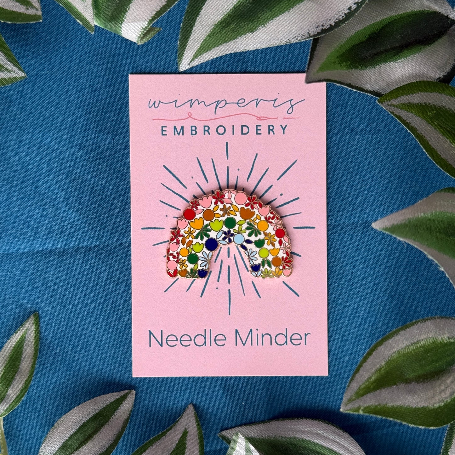 Wimperis Embroidery Needle Minder with floral rainbow design on pink card, surrounded by green leaves on blue fabric background.