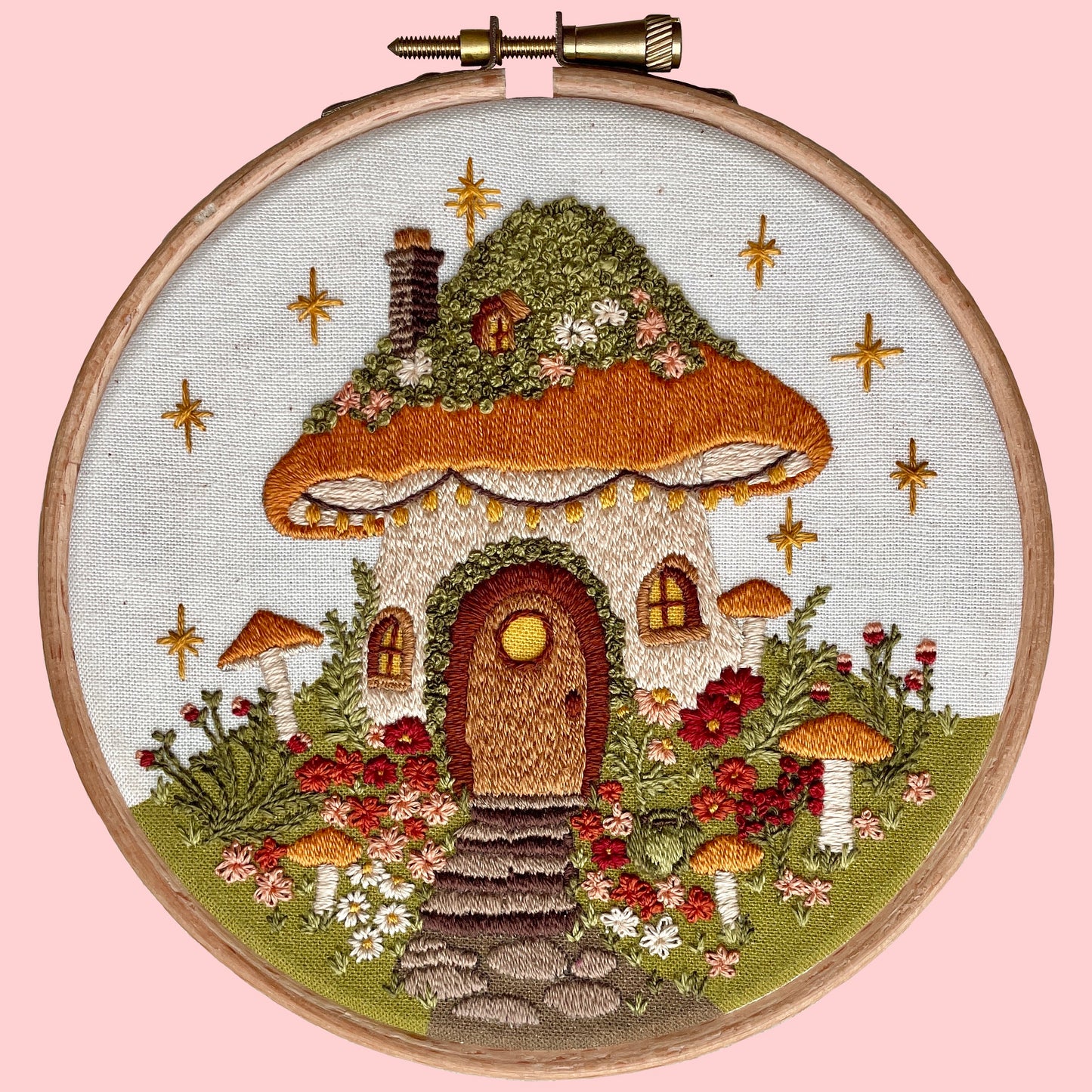 Embroidery kit depicting a whimsical mushroom house with a printed background, surrounded by flowers and mushrooms.