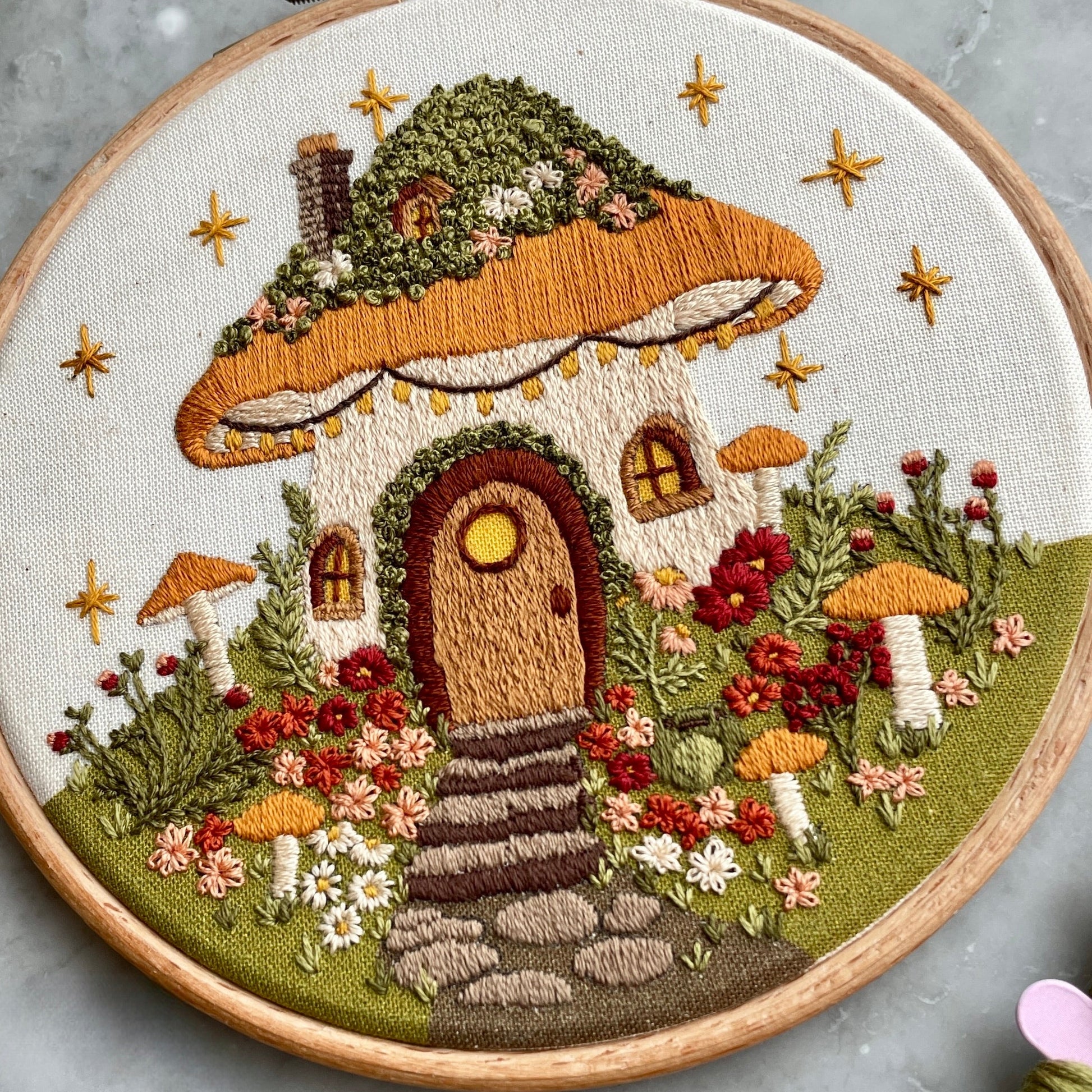 Embroidery of a mushroom house with flowers and stars in a wooden hoop.