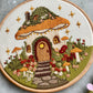 Embroidery of a mushroom house with flowers and stars in a wooden hoop.
