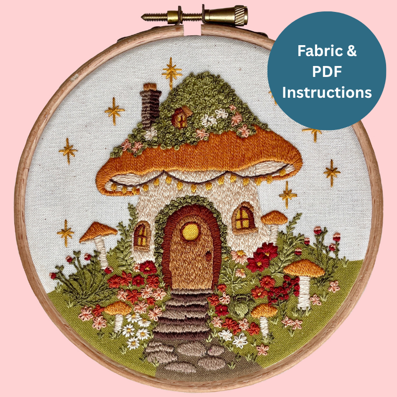 Embroidery hoop with a mushroom house design on fabric, against a pink background.