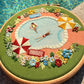 Embroidery hoop with a pool scene featuring people, umbrellas, and flowers, placed over a real pool.