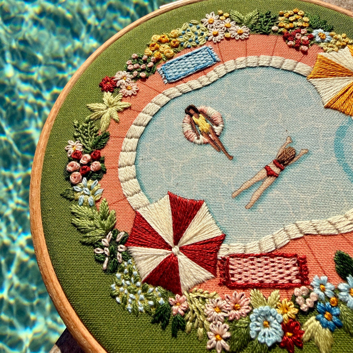 Embroidery hoop with a pool scene design featuring people by a pool, flowers, and umbrellas.