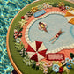 Embroidery hoop with a pool scene design featuring people by a pool, flowers, and umbrellas.