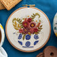 Embroidery hoop with floral design on a blue surface with a cup of coffee, cookies, and teapot.