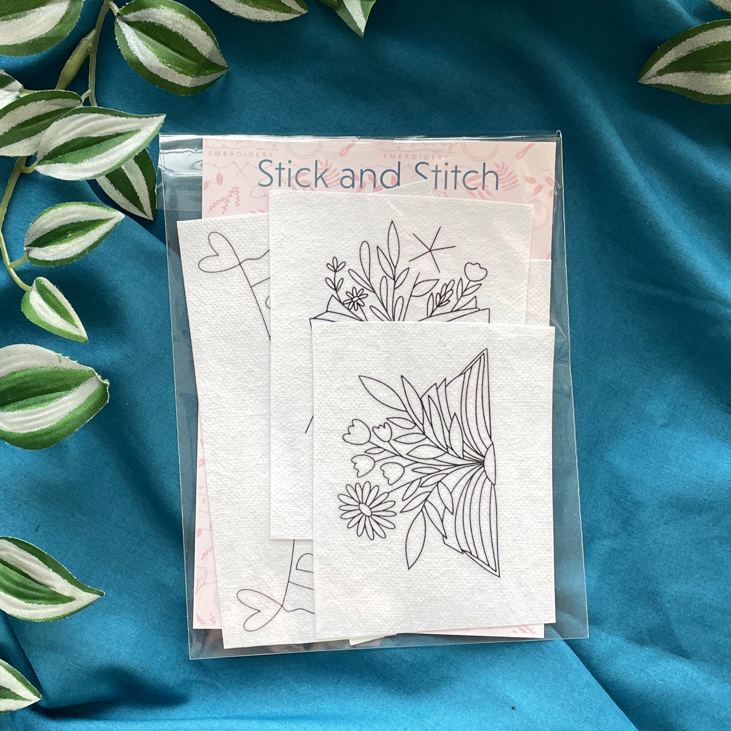 Packaging of Stick and Stitch embroidery kit placed on a blue fabric with leaf accents, displaying the brand name and contents of the package.