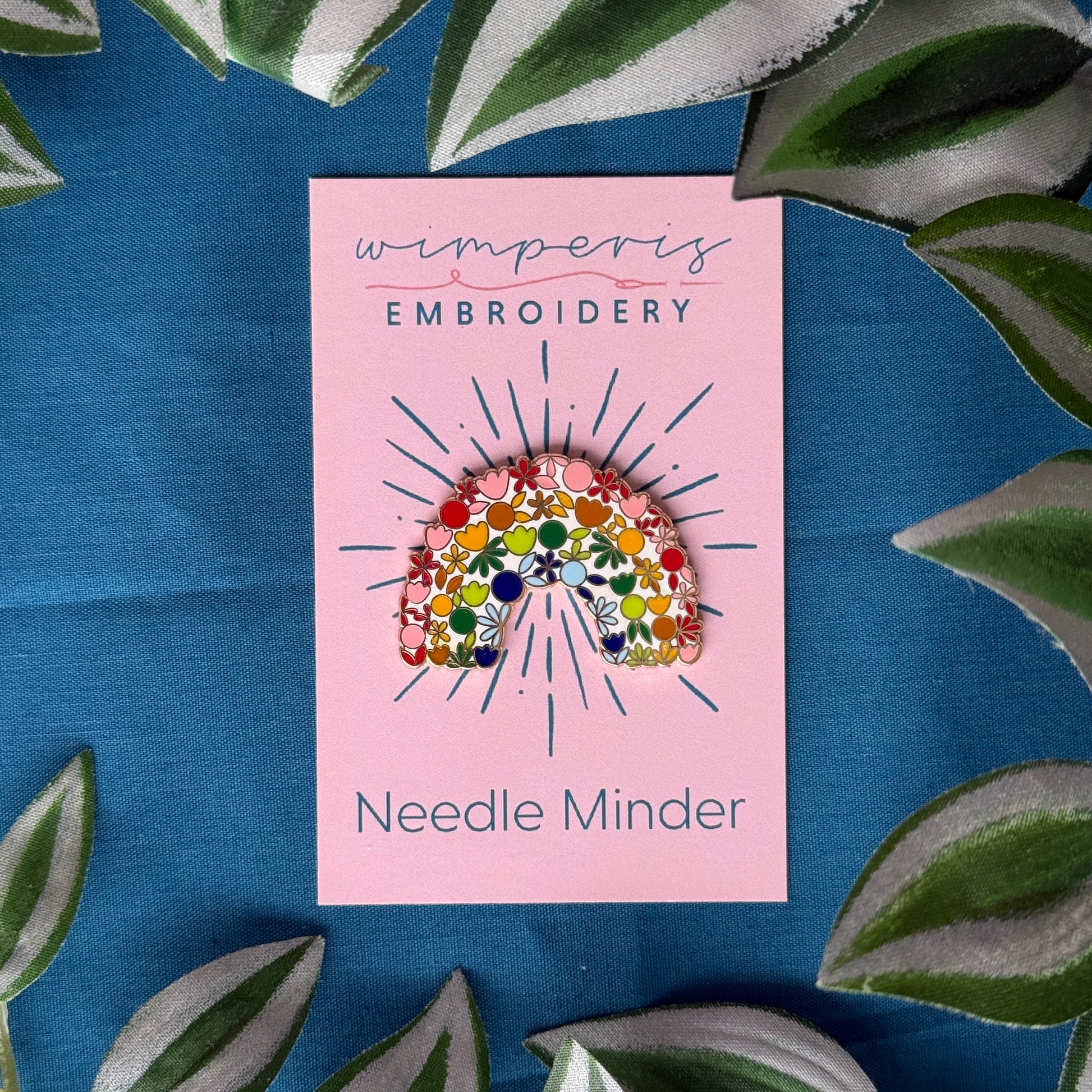Wimperis Embroidery Needle Minder with floral rainbow design on pink card, surrounded by green leaves on blue fabric background.