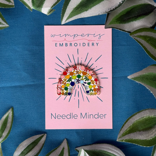 Wimperis Embroidery Needle Minder with floral rainbow design on pink card, surrounded by green leaves on blue fabric background.