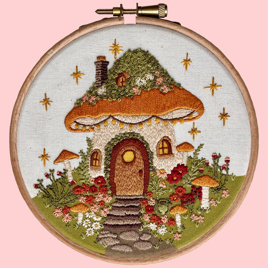 Embroidery kit depicting a whimsical mushroom house with a printed background, surrounded by flowers and mushrooms.