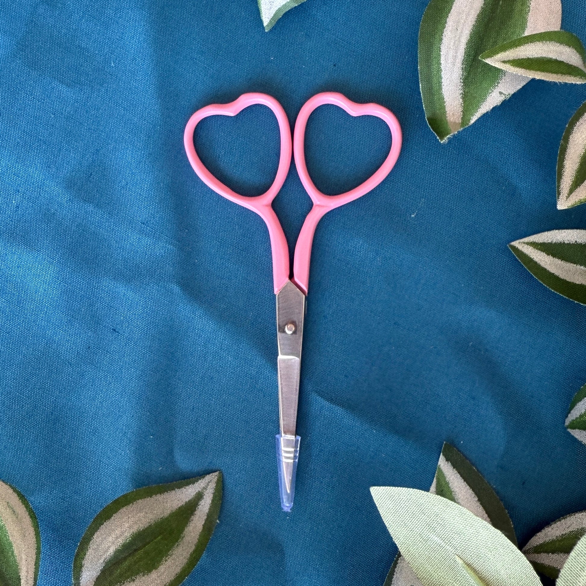 Pink scissors with heart-shaped handles on a blue fabric background with leaf patterns